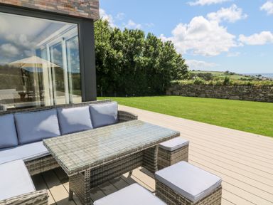 An outdoor patio with a glass table and cushioned wicker chairs next to a house with large windows and a green lawn at Atlantic View in Germoe