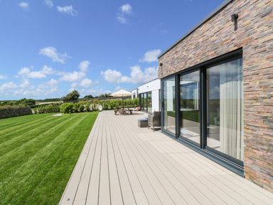 A house with large sliding glass doors opening onto a wooden deck with outdoor furniture and a grassy lawn at Atlantic View in Germoe
