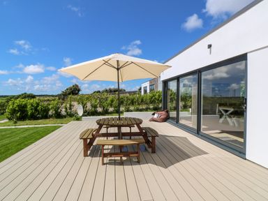 An outdoor deck with a wooden picnic table and beige umbrella next to large glass sliding doors at Atlantic View in Germoe