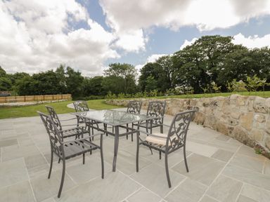 An outdoor patio with a glass table and six metal chairs near a stone wall and greenery at The Corn Store in St Stephen