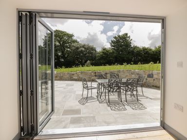 An open patio door leading to a stone terrace with a metal table and six chairs surrounded by a stone wall and trees at The Corn Store in St Stephen