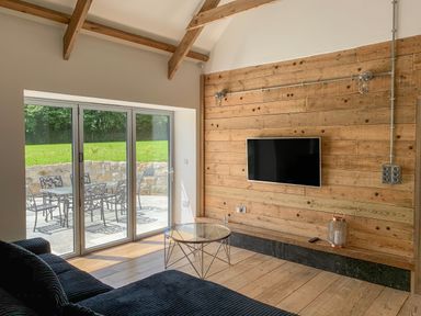 A living room with wooden walls and ceiling beams a glass coffee table and a sofa facing a wall-mounted television at The Corn Store in St Stephen