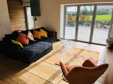 A living room with a black sofa with multi-colored cushions a brown armchair a glass coffee table and a patterned rug in front of large sliding glass doors at The Corn Store in St Stephen