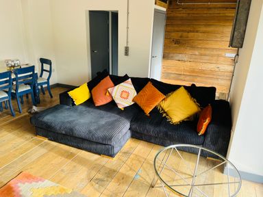 A living room with a black sectional sofa with colorful cushions a glass round coffee table wooden floor and a dining table with blue chairs at The Corn Store in St Stephen