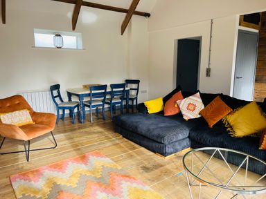 A living room with a dark sectional sofa with colorful cushions a blue dining table with chairs an orange armchair and a patterned rug on wooden floors at The Corn Store in St Stephen