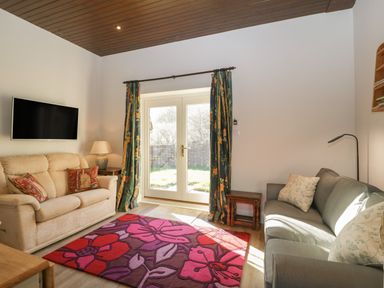 A living room with two sofas a wall mounted television a floral area rug and glass doors with curtains at Stable Cottage in Triscombe