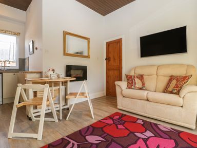 A living room with a beige sofa and two red pillows a wall mounted TV a wooden door a small wooden table with chairs a floral rug a mirror and a kitchen area at Stable Cottage in Triscombe