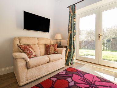 A beige sofa with patterned cushions beside a lamp on a side table and a wall-mounted TV near glass doors with floral curtains and a colorful rug on the floor at Stable Cottage in Triscombe