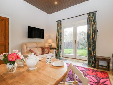 A living room with a sofa TV table lamp dining table flowers teapot plates and glass doors with curtains at Stable Cottage in Triscombe