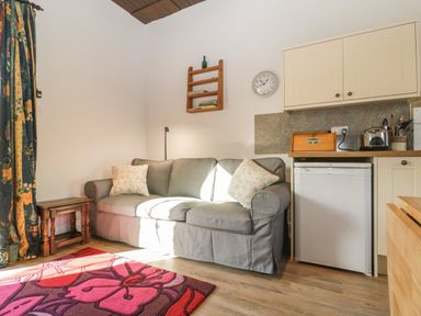 A gray sofa with two pillows next to a small wooden table and kitchen cabinets with a mini fridge at Stable Cottage in Triscombe