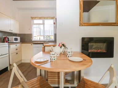 A kitchen with a wooden dining table set with plates and mugs next to a wall-mounted electric fireplace at Stable Cottage in Triscombe