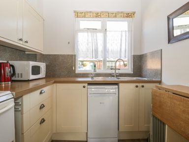 A kitchen with beige cabinets a white microwave red kettle a dishwasher under the sink and a window with curtains at Stable Cottage in Triscombe