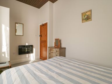 A bedroom with a striped bedspread a wooden door a small dresser with a mirror and a wall-mounted heater at Stable Cottage in Triscombe