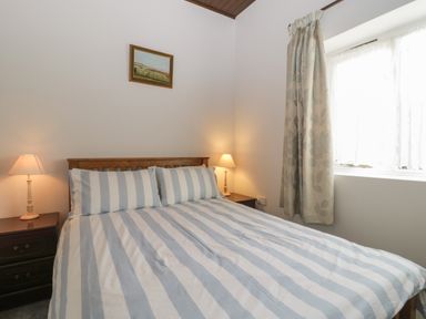 A bedroom with a bed with striped bedding two bedside tables with lamps a window with curtains and a painting on the wall at Stable Cottage in Triscombe