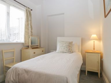A bedroom with a single bed with a pillow a bedside table with a lamp a chair a dresser with a mirror and a window with curtains at Stable Cottage in Triscombe