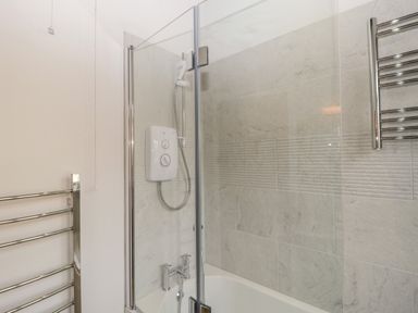 A bathroom with a bathtub shower combination a glass shower screen and a wall-mounted electric shower at Stable Cottage in Triscombe