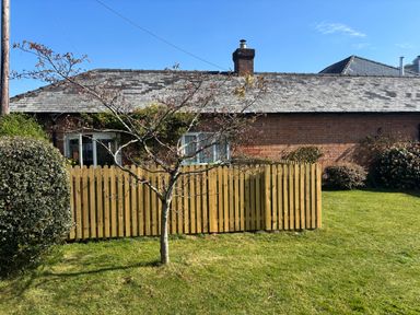 A small tree and bushes in front of a wooden fence with a brick house behind at Stable Cottage in Triscombe