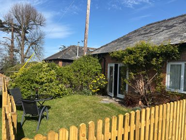 A small fenced garden with two black chairs and a table with a bottle of wine in front of a brick house at Stable Cottage in Triscombe