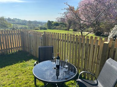 A garden with a round glass table, two chairs, a bottle of wine, and two wine glasses by a wooden fence and a blooming tree at Stable Cottage in Triscombe