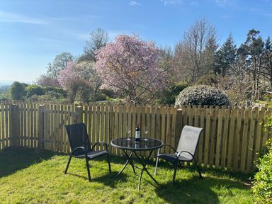 A garden with two chairs and a round table with a bottle and two glasses near a wooden fence and trees at Stable Cottage in Triscombe