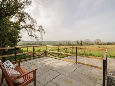 A stone patio with a wooden bench and a railing overlooking a grassy field with sheep at The Wash House in Sibford Ferris