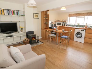 An open plan living room and kitchen with a sofa wood stove dining table and chairs and shelves with books at The Wash House in Sibford Ferris
