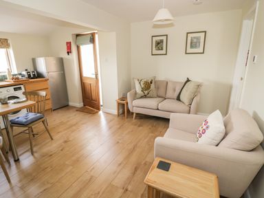 A living area with two sofas, a wooden door, framed pictures on the wall, and a small kitchen with appliances at The Wash House in Sibford Ferris