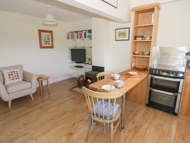 A kitchen and living area with a wooden dining table, chairs, a cake, a stove, an armchair, shelves with bowls, a TV, a wood burner, and framed pictures at The Wash House in Sibford Ferris