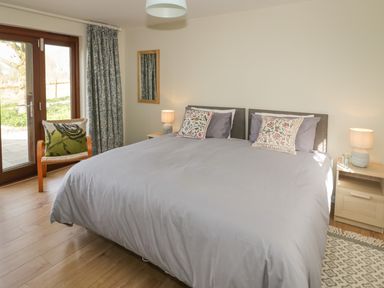 A bedroom with a large bed two bedside tables with lamps a chair with a cushion patterned curtains glass door and a wall mirror at The Wash House in Sibford Ferris