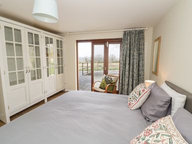 A bedroom with a large bed, pillows, a wooden chair with cushions, glass-paneled wardrobes, and a window with patterned curtains at The Wash House in Sibford Ferris