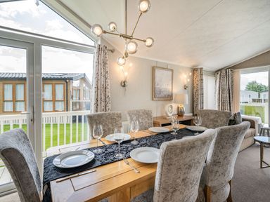 A dining area with a wooden table set for six and large windows showing outdoor buildings at Links Lodge in Flookburgh