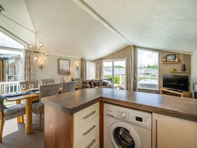 A kitchen counter with a washing machine a dining table with chairs and a living area with a television at Links Lodge in Flookburgh