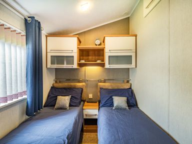 A bedroom with two single beds side by side with blue bedding and pillows two white overhead cabinets and a small shelf with a clock in the center at Links Lodge in Flookburgh