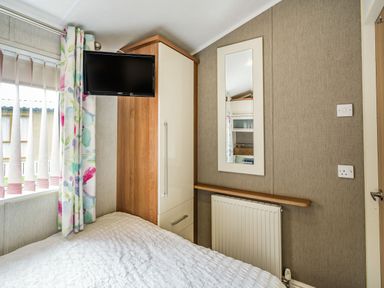 A bedroom corner with a mounted tv wardrobe a wall mirror radiator window with floral curtains at Links Lodge in Flookburgh