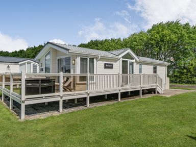 A white single-story mobile home with large windows and a raised deck with outdoor furniture on a lawn at Links Lodge in Flookburgh