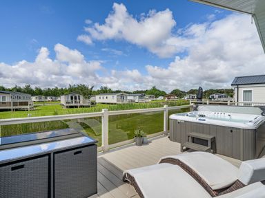 A deck with a hot tub two lounge chairs and a storage cabinet overlooking a grassy area with multiple mobile homes at Links Lodge in Flookburgh
