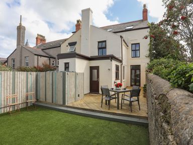 An outdoor patio area with a table and four chairs on wooden decking next to a cream colored house with dark framed windows and doors and a lawn with two wooden chairs at Monfa in Cemaes Bay