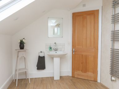 A bathroom with a pedestal sink a mirror a wooden door a towel holder with a dark towel a small table with a vase of flowers and a heated towel rack at Monfa in Cemaes Bay