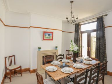 A dining room with a wooden table set with white dishes and glasses a vase with flowers on the table a fireplace with two small plants a painting above it two chairs to the side and a chandelier above at Monfa in Cemaes Bay