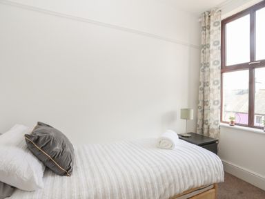 A bedroom with a single bed with white bedding and a gray pillow a black nightstand with a lamp and two small plants on the windowsill with floral curtains at Monfa in Cemaes Bay