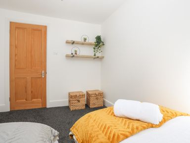 A bedroom corner with wooden door two wicker storage boxes floating shelves with plants and a bed with white towel and orange blanket at Monfa in Cemaes Bay
