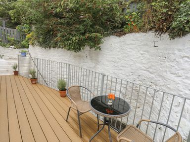 An outdoor patio with a round glass table and two wicker chairs near a white stone wall with plants at Glan Meon Cottage in Barmouth
