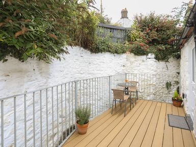 An outdoor patio with wooden flooring metal railing a small table with two chairs potted plants and a white stone wall at Glan Meon Cottage in Barmouth