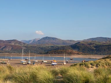 A coastal scene with boats on a sandy shore surrounded by grassy dunes and mountains in the background at Glan Meon Cottage in Barmouth