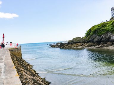 A coastal walkway with people near a small lighthouse and rocky shoreline at Bay Tree Cottage in West Looe