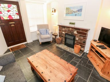A living room with a wooden coffee table a fireplace a gray armchair a wooden TV stand and a door with stained glass at Bay Tree Cottage in West Looe