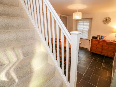 A carpeted staircase next to a dining room with a wooden table chairs sideboard clock and lamp at Bay Tree Cottage in West Looe