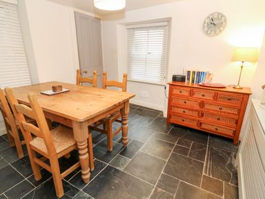 A dining area with wooden table and chairs a chest of drawers with books clock on the wall and a lamp at Bay Tree Cottage in West Looe