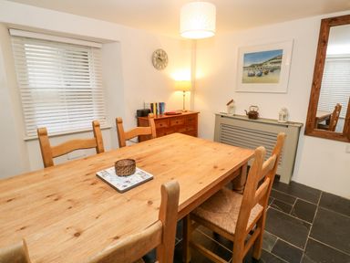 A dining room with a wooden table and chairs a sideboard with books and a lamp a wall clock a framed picture and a large mirror at Bay Tree Cottage in West Looe
