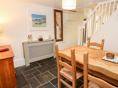 A dining area with a wooden table and chairs near a staircase and a radiator cover with decorative items at Bay Tree Cottage in West Looe